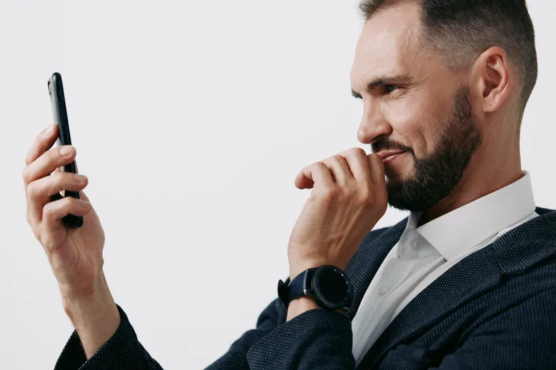 A professional business man sits against an isolated white background, looking at his smartphone with a thoughtful, confident expression, signaling focus and achievement.