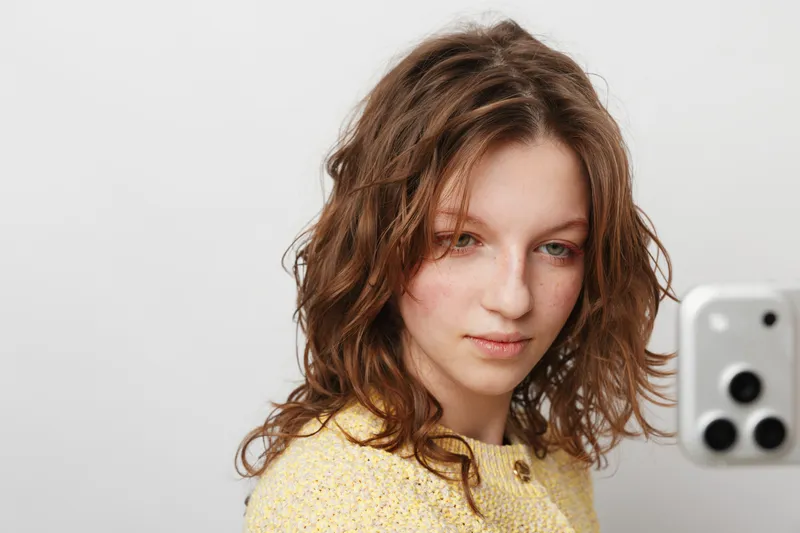 Young woman with curly hair wearing yellow sweater takes selfie with smartphone on white background. Natural light highlights her relaxed expression and casual style. Concept of social media.