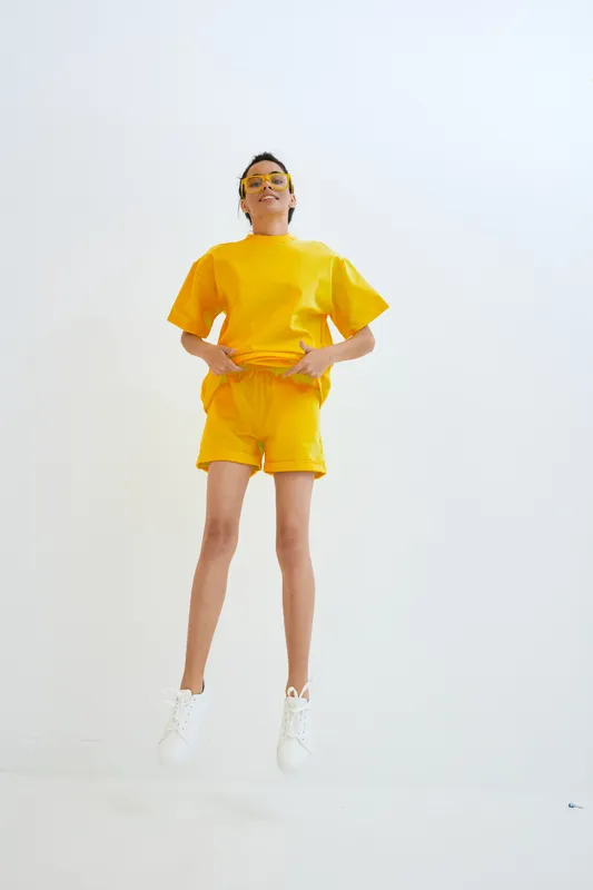 young woman in yellow activewear jumping against white background with sunglasses, casual style, energetic pose, minimalistic bright setting