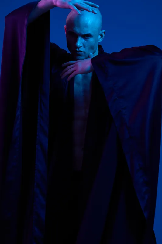 Mysterious man in dark clothing elegantly posing against a vivid blue background. The image captures an intriguing blend of shadows and lighting effects.