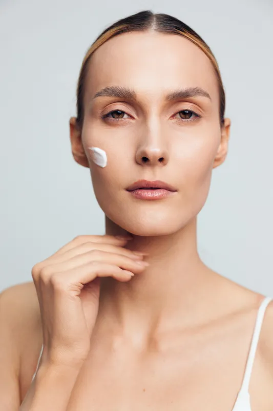 A young woman with flawless skin applies gentle cream to her cheek, concentrating on skincare routine, standing in a bright minimal studio with soft lighting and a neutral backdrop, isolated.