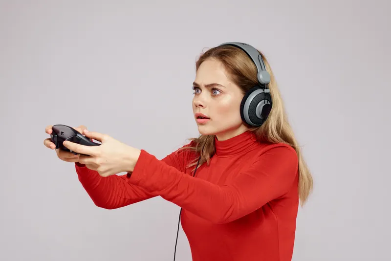 Женщина в наушниках, с джойстиком в руках, играет в игры, весёлый досуг, образ жизни, развлечения, красная рубашка, серый фон