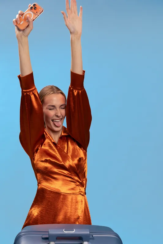 Excited woman in shiny orange dress holds phone above head, smiling with closed eyes, standing behind suitcase on blue background. Concept of travel, joy, and celebration.