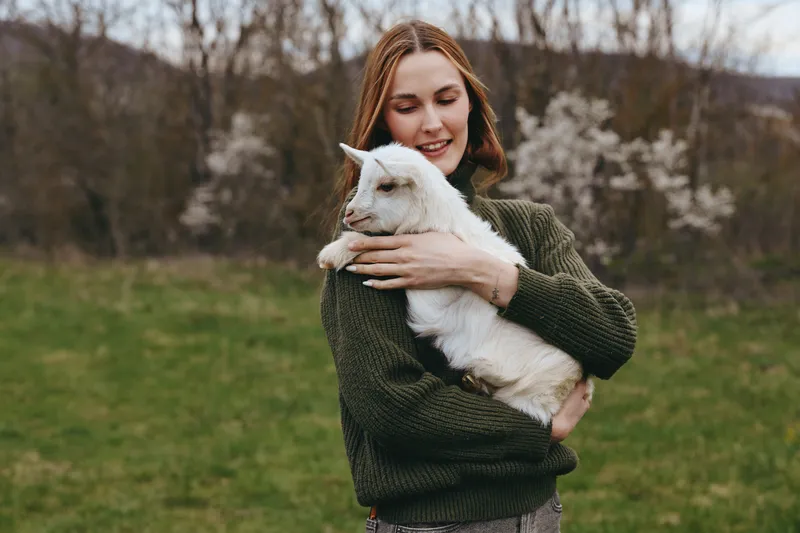 Young woman in a green sweater gently holds a white baby goat outdoors during spring, enjoying a peaceful moment in nature with a soft smile on her face.