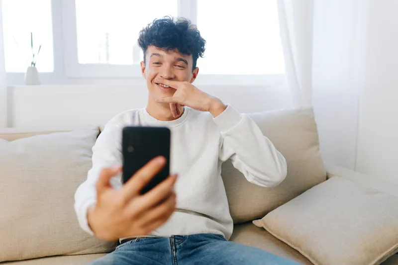 Счастливый молодой человек улыбается и позирует для селфи на своём смартфоне, удобно сидя на светлом диване в спокойной, хорошо освещённой комнате