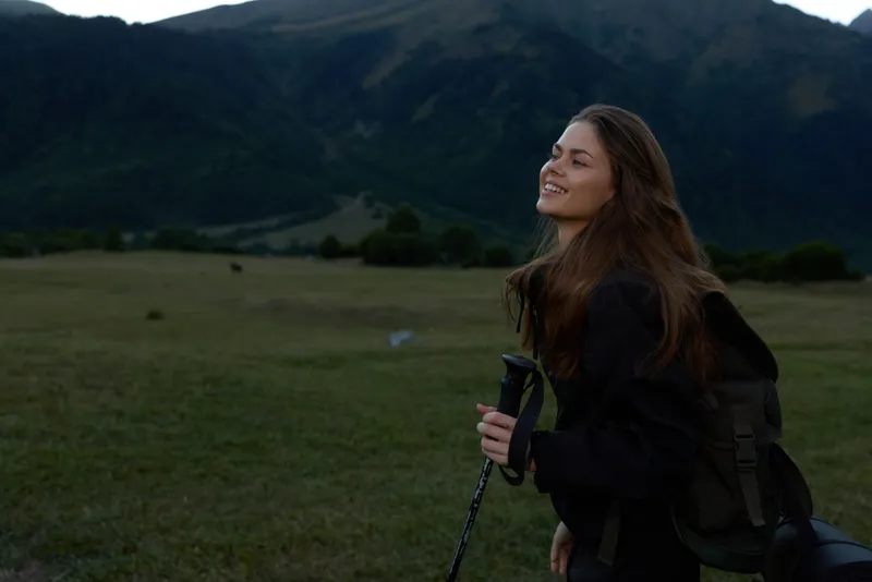 A smiling hiker woman stands in a wide meadow with rolling hills and distant mountains. She wears a backpack and holds trekking poles, enjoying nature and fresh air.