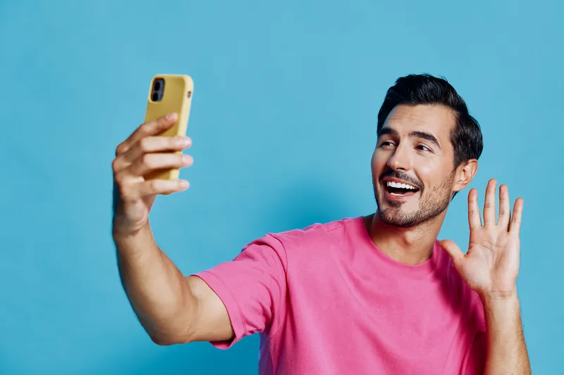 Full body smiling cheerful young man wearing pink t-shirt looking at camera while waving and using mobile phone isolated on solid blue background. People lifestyle concept.
