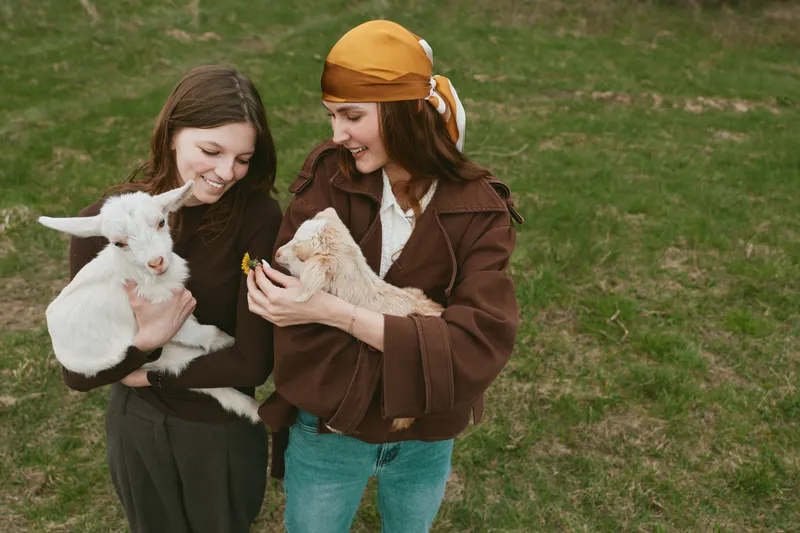 Two women in casual clothes smile while holding a goat kid, sharing a playful outdoor moment with gentle animals, warm interaction, rustic park setting, lifestyle scene with affection and friendship