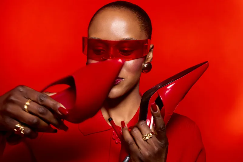 Elegant dark skinned African woman with shaved head holds shiny red high heels, wearing red clothes and translucent red glasses. Concept for fashion, style, and modern beauty portrait.