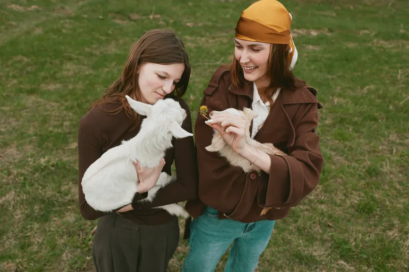 Two young women enjoy a spring day outdoors as they hold and feed a white baby goat in a grassy field, sharing a joyful and natural lifestyle moment together.