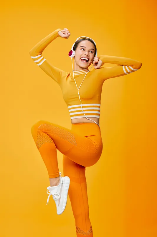 Bright studio scene with a cheerful woman in orange athletic wear jumping mid-dance, wearing headphones, showcasing fitness energy, sport movement and vibrant lifestyle in a warm yellow backdrop.