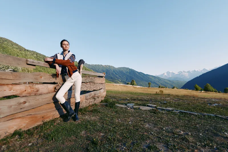 Woman leaning on wooden fence in countryside meadow with distant mountains, wearing riding boots and casual jacket, portrait lifestyle outdoors in wide open field under clear sky.