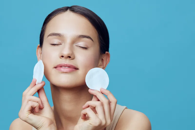 Young woman with a serene expression holding cotton pads for skincare on a blue background, representing beauty and self care