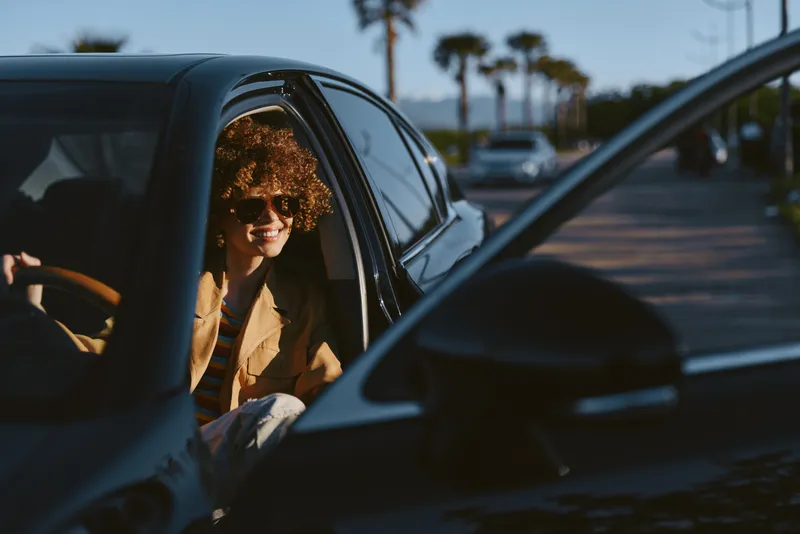 Cheerful woman in a car, sunglasses on, sunlight streaming through the window, relaxed pose, warm retro tones, natural colors, outdoor street scenery behind
