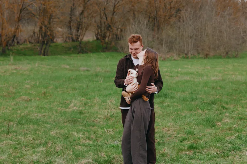 A couple outdoors in a grassy field gently embracing a young goat kid, smiling as a warm lifestyle moment. The scene conveys affection, family bonding, and care for animals in a bright, open