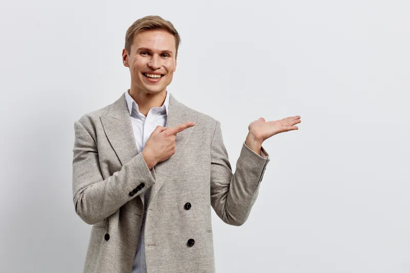 Cheerful young man in gray blazer smiling and pointing to empty space against light gray background, showing a presentation or product. Casual business style, studio indoor shot, lifestyle concept, po