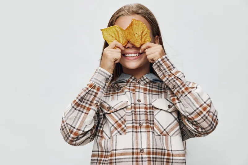 Child girl smiling and covering eyes with autumn leaves in plaid shirt, standing against white background for seasonal fun and fall concept.