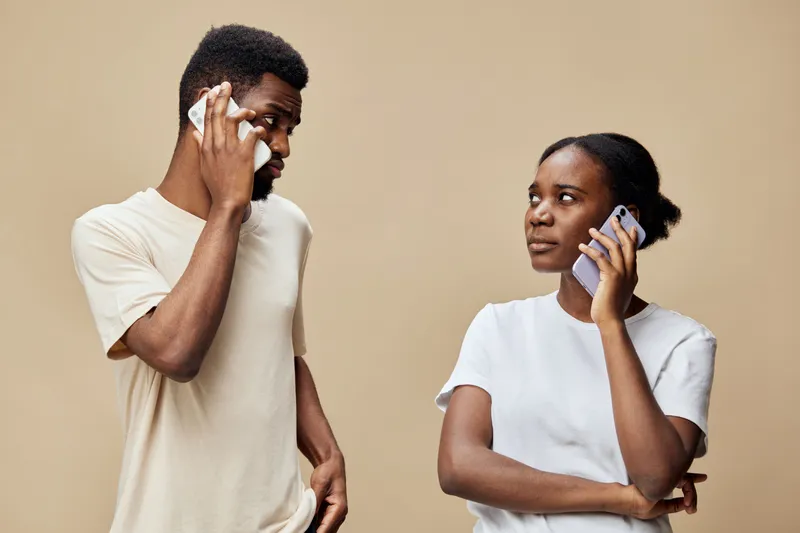 Young Black couple communicating via smartphones on a neutral background, expressing emotions in a casual, modern setting with contemporary clothing