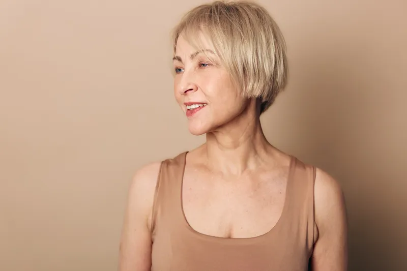 Portrait of confident mature woman with short blonde hair smiling softly against beige background. Concept for aging beauty, natural look, self confidence, and minimalism in lifestyle.