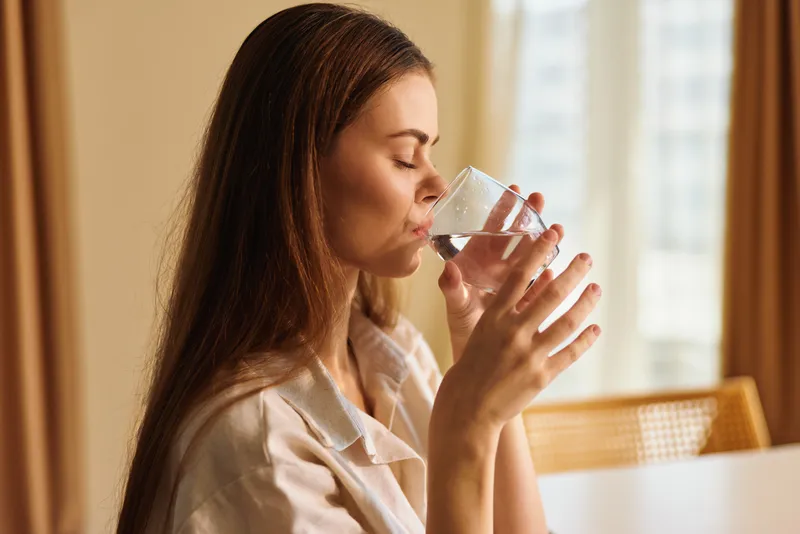 Woman drinking water from a glass at home, portrait profile with closed eyes and long hair, morning light through window, relaxed hydration routine for calm wellness.