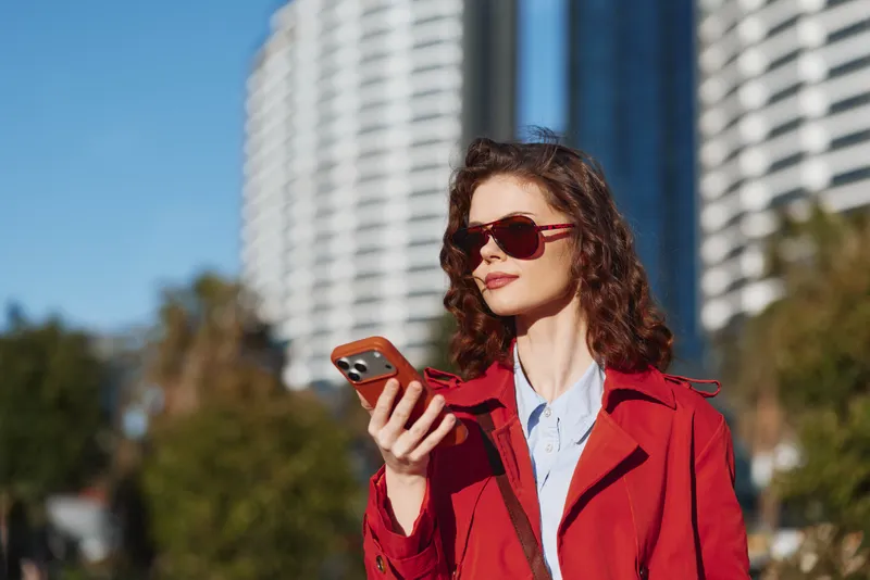 Young woman with curly hair wearing a red coat checks her smartphone outdoors with modern buildings in the background. It conveys confidence and urban style.