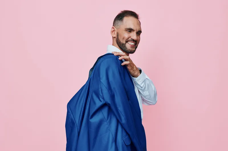 A confident man in a professional stance carries a vibrant blue jacket against a soft pink background, creating an isolated color field that highlights modern business mood and strong emotional