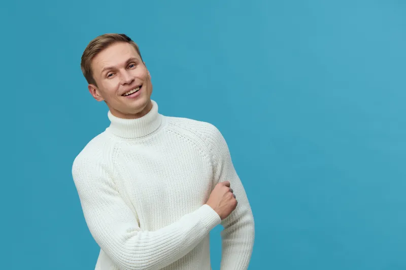 Smiling man wearing white knit turtleneck sweater standing against a bright blue background, casual style, cheerful expression, studio portrait, fashion concept, lifestyle.