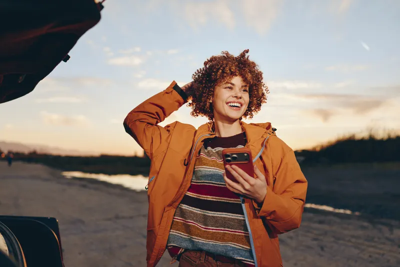 Woman lifestyle outdoors at beach sunset, smiling woman with curly hair, wearing orange jacket, laughing while holding smartphone, warm evening light, casual travel moment