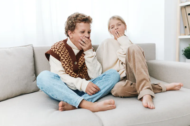 Two children sit on a light sofa, sharing a shy laugh while covering mouths, wearing cozy sweaters, in a bright room, creating a candid, wholesome moment of playful secrecy.