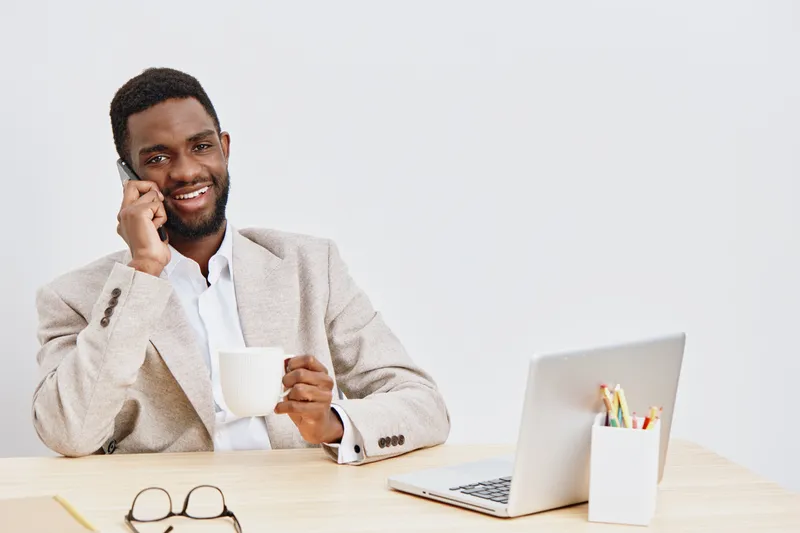 Smiling man talking on mobile phone while holding coffee mug sitting at a desk with laptop and glasses on a plain white background. Business lifestyle concept