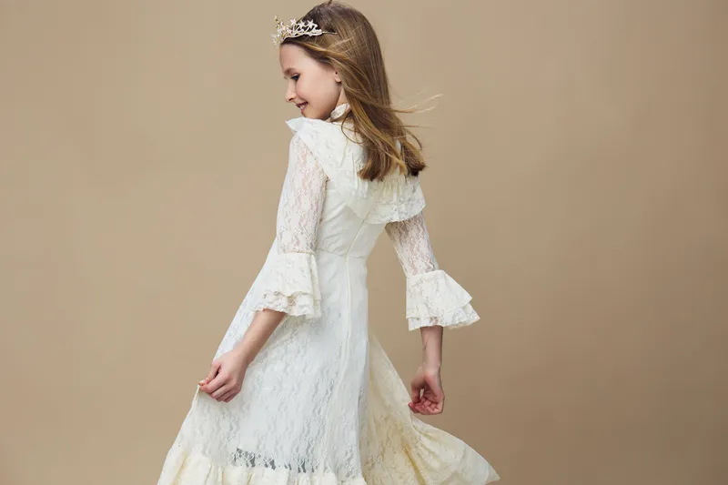 Young girl in elegant white lace dress with bell sleeves, expressing joy, against a soft beige background, perfect for children s fashion and portrait photography