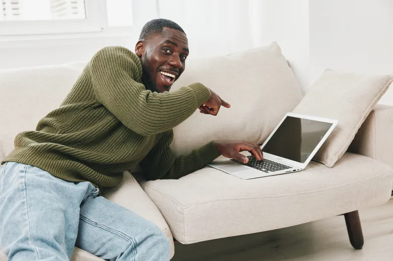 Happy African American man using a laptop on a cozy couch, showcasing a vibrant atmosphere of productivity and relaxation in a modern interior