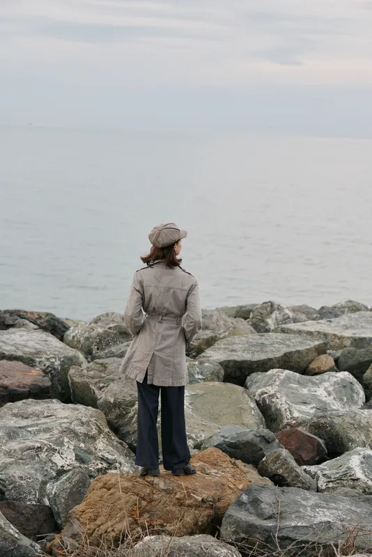 Woman in coat and hat stands on rocks looking at calm sea under cloudy sky. Concept of solitude, nature, and peaceful coastal landscape in cool weather.