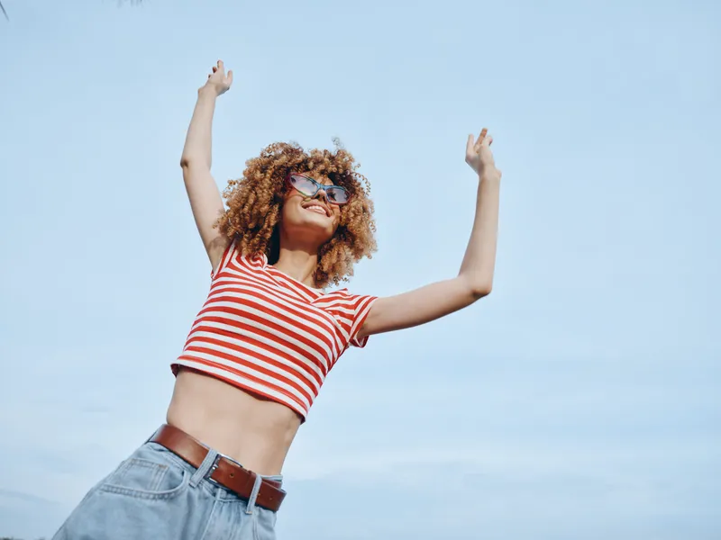 A joyful person with curly hair wearing a striped red and white top and denim jeans stands against a clear blue sky, arms raised in celebration.