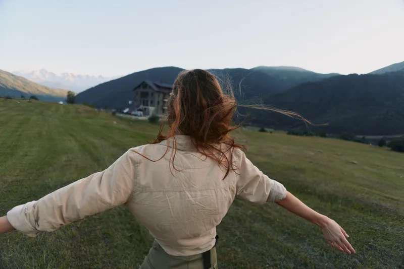 Woman with long hair wearing beige jacket walking on green grass in mountain landscape. Concept of freedom, lifestyle, and outdoor adventure during daytime.