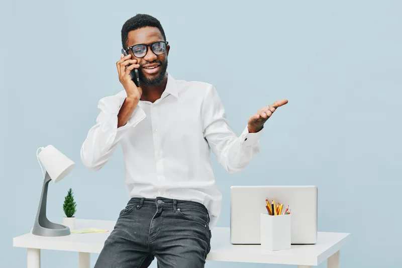 Happy young African American man in casual white shirt and jeans talking on mobile phone indoors, standing next to desk with laptop and stationery, cheerful expression, bright mood, modern workspace, 