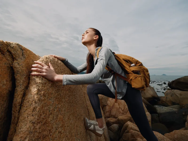 Young woman climbing rocky terrain with determination, wearing sportswear and a backpack, conveying adventure and resilience in a natural setting