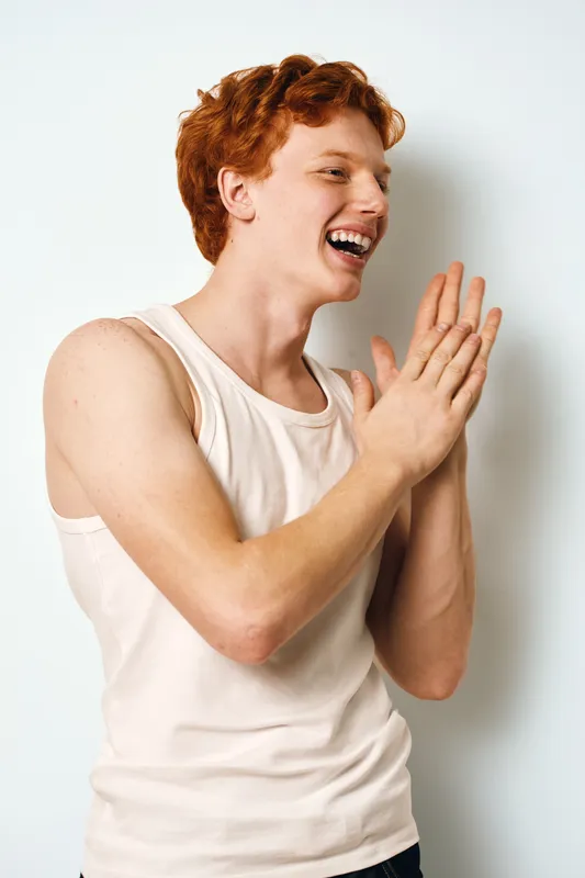 Young man with red hair wearing white tank top laughing and clapping hands against plain white background. Casual smiling male in sleeveless shirt in joyful pose indoors.