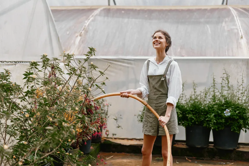Happy female gardener with a hoop in a greenhouse, wearing a casual outfit and smiling, surrounded by lush plants and a bright atmosphere