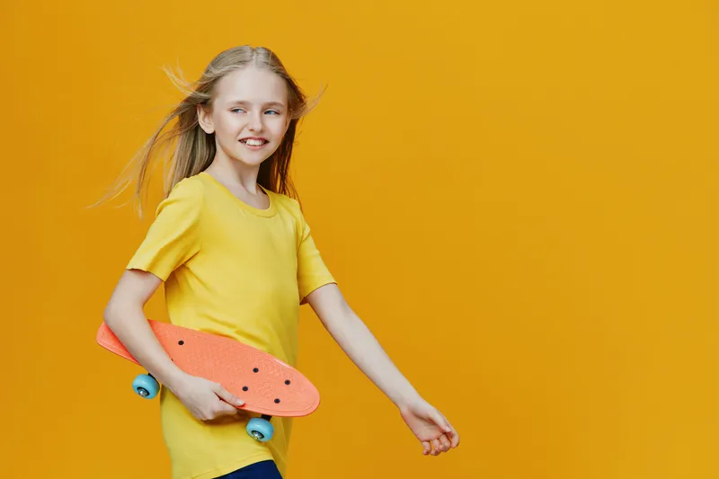 Young girl with blonde hair in a yellow t shirt, joyfully walking with a skateboard against a bright yellow background Fun, playful, and energetic youth concept