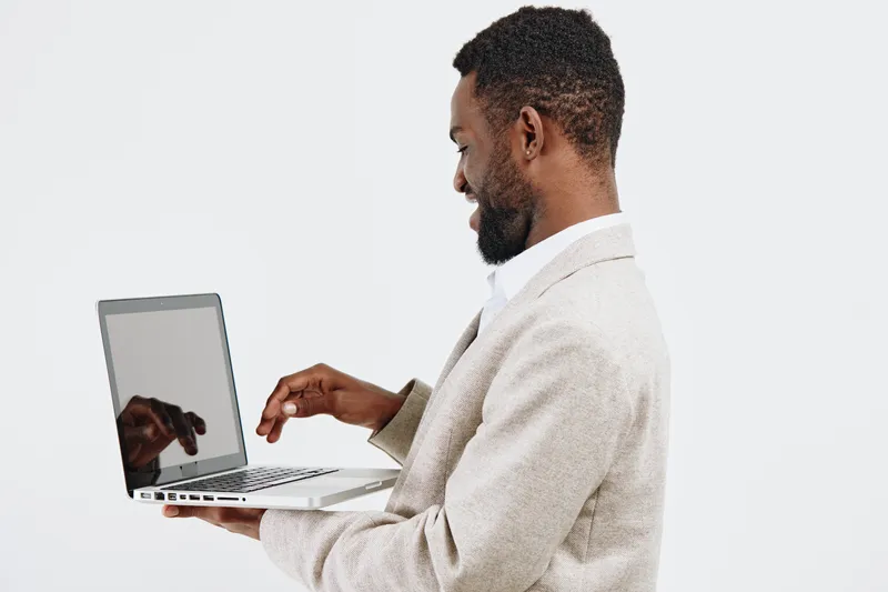 Professional smiling man with dark skin and beard, wearing beige blazer, working with silver laptop, isolated on plain light grey background, studio portrait, technology and business concept.