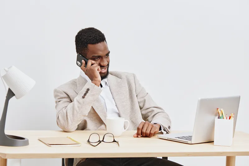 Professional young African American man working in office, using laptop and talking on mobile phone, wearing beige blazer, glasses and white shirt, in modern workplace environment, lifestyle and busin