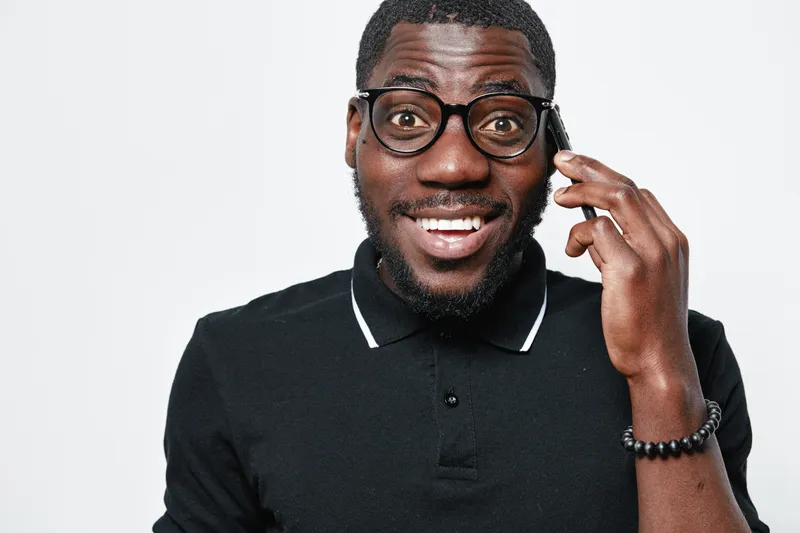 African American man with dark skin wearing glasses and black polo shirt talking on smartphone with happy expression isolated on white background. Communication, technology and casual style.