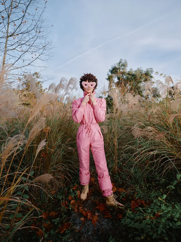 A person in a pink jumpsuit and masquerade mask stands in a tall grass field, posing with hands near the face for a creative fashion portrait in natural autumn light.