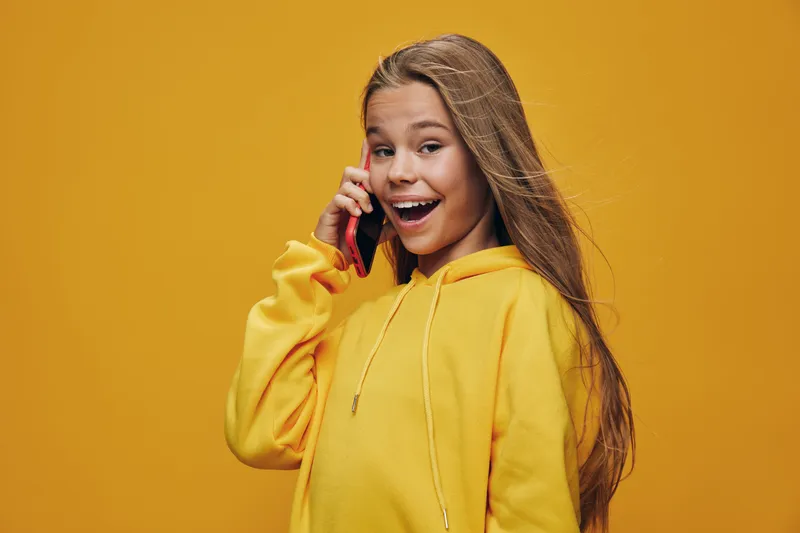 Girl wearing yellow hoodie smiling and talking on smartphone over yellow background. Young female with long hair expressing happiness and communication concept in studio.