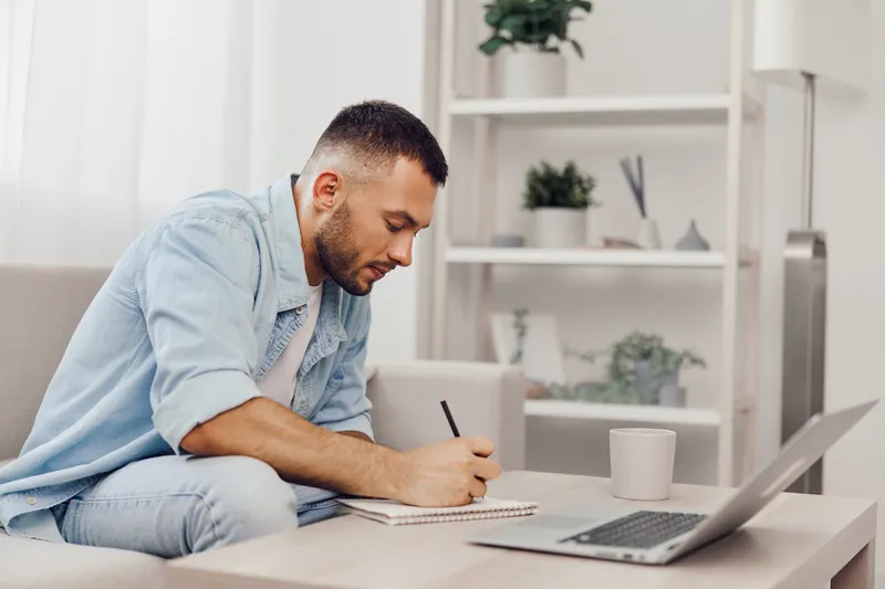 Focused man writing notes at home, sitting at a table with a laptop and a cup, showcasing productivity and modern living in a minimalist style.