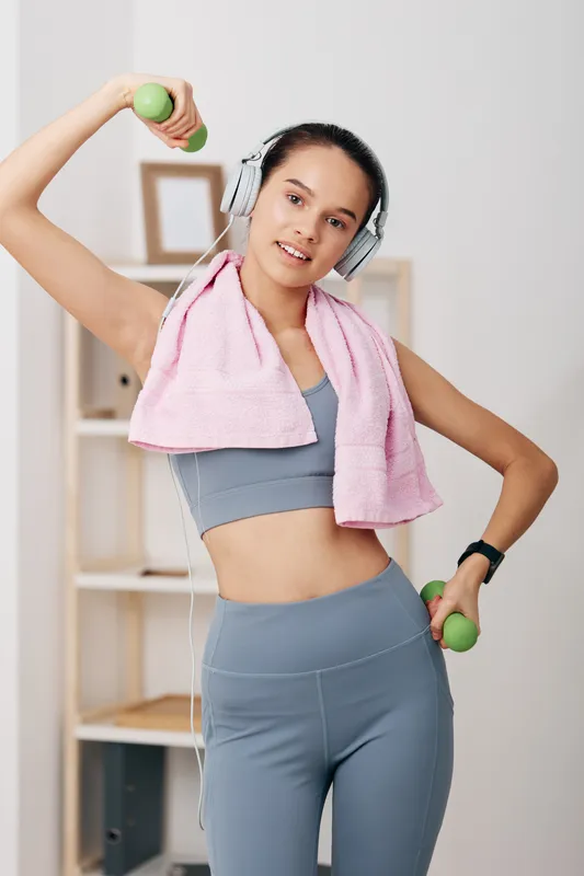 Fitness scene of a woman in athletic wear lifting dumbbells at home, headphones on, pink towel over shoulders, capturing dynamic movement, motivation, and active sport vibe.