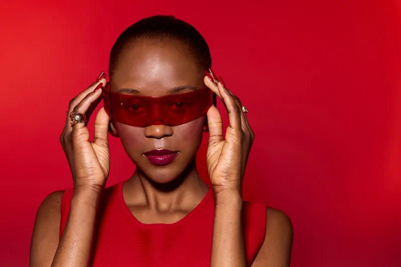 Confident African woman with dark skin wearing red sleeveless dress and red sunglasses adjusts eyewear on red background. Concept for fashion, style, and modern beauty portrait.