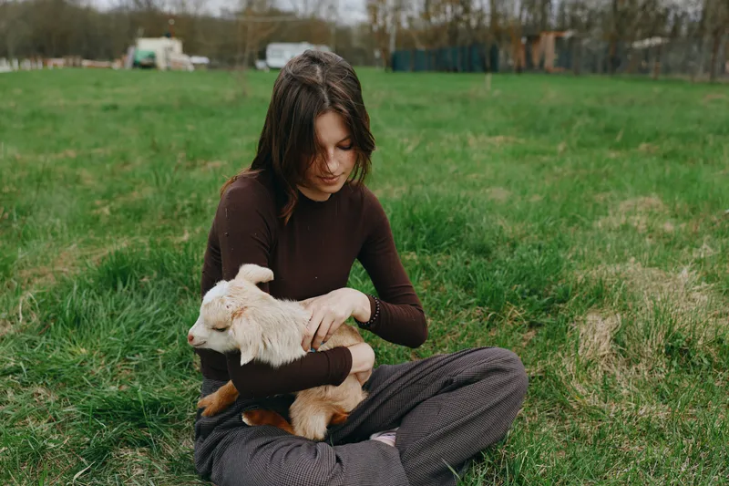 Casual woman outdoors, seated on grass, cradling a gentle goat kid in her arms, warm sunlight, calm interaction, earthy tones, tranquil countryside scene, lifestyle portrait with animal companion