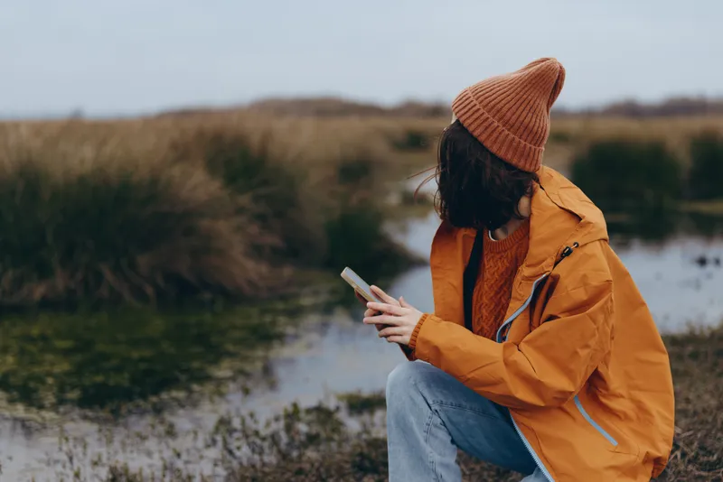 Woman lifestyle outdoors by river, orange jacket and knit beanie, seated on grassy bank, looking at smartphone, thoughtful relaxed mood, soft cloudy light, natural colors, calm nature